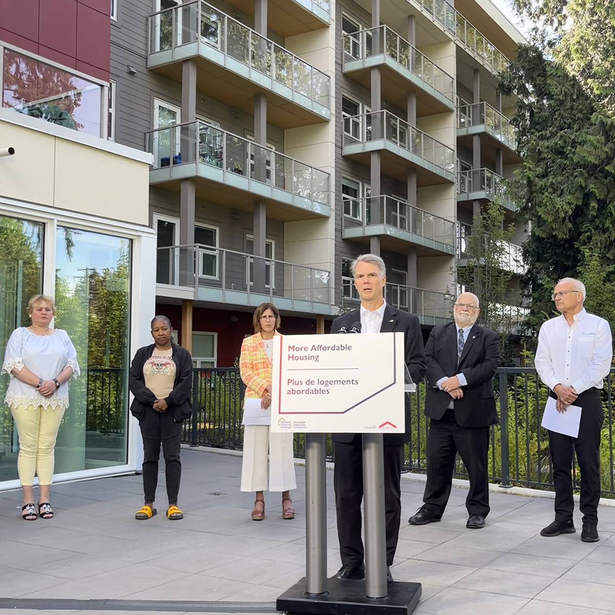 A man speaks at a podium with a "More Affordable Housing" sign at a housing announcement