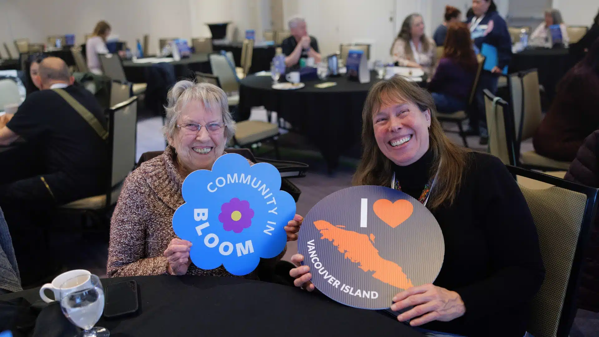 Two women holding "Community in Bloom" and "I love Vancouver Island" signs at a meeting